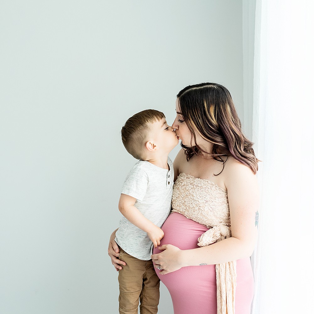 In Studio + Outdoor Maternity Photography and Photo Session Warwick, RI, Conimicut Village, Conimicut Point - mother and son standing near window embracing with a hug and kissing
